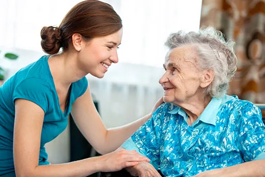 A smiling support worker kneeling beside an older woman and holding her hands in her own home