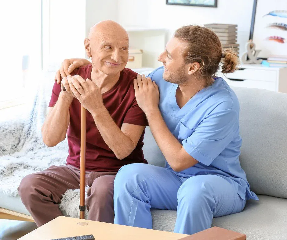 A Pamis Care support worker in blue scrubs sitting on a sofa with an older man, sharing a smile in his living room