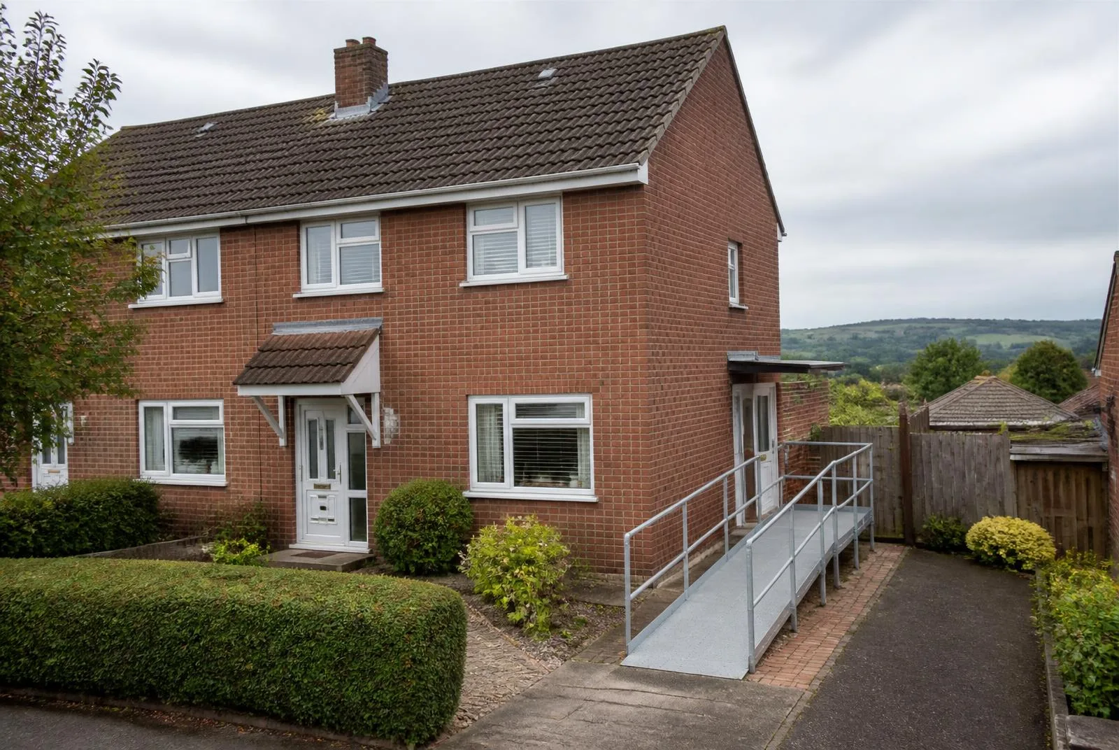 Representative red-brick semi-detached supported-living home in Hereford, with a level ramped side entrance, handrail, white UPVC windows, small front garden and a staff parking bay